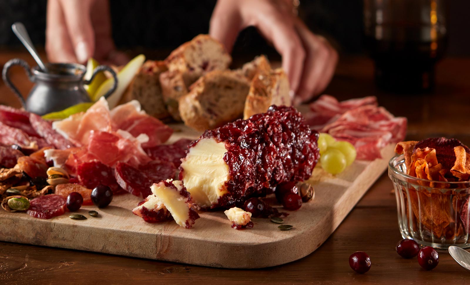 A pair of hands preparing a wooden board with cheddar cheese with cranberry log, assorted sliced meats, fruits and breads.