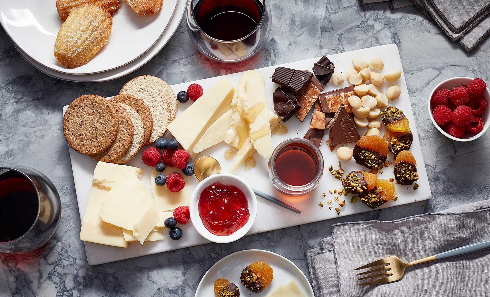 A charcuterie dessert board filled with slices of cheddar cheese, dark chocolate, nuts, jam, blueberries, raspberries and crackers.