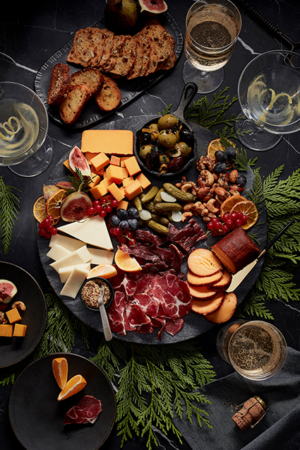A winter inspired charcuterie board with a chipotle seasoned cheese log, cheddar cheese cubes, sweet pickles, mixed raw nuts, orange slices, grapes, blueberries and sliced meat.